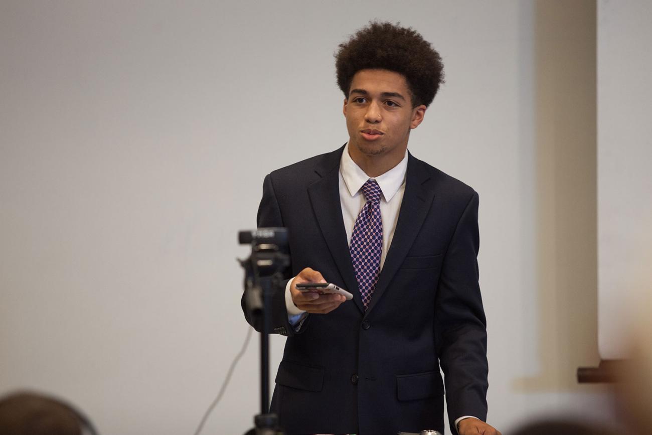 Young man in blue suit and white shirt holding up phone in front of class