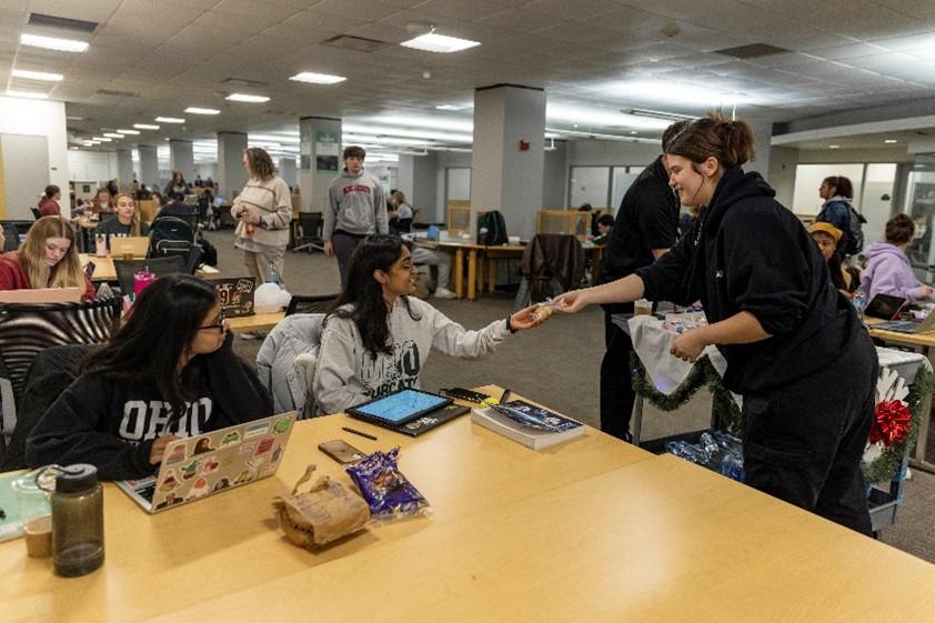 A young woman hands a packaged cookie to a student seated at a table in a crowded study area