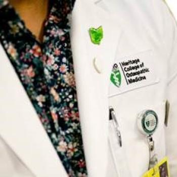 A person wearing a white coat with the Heritage College of Osteopathic Medicine logo on it.