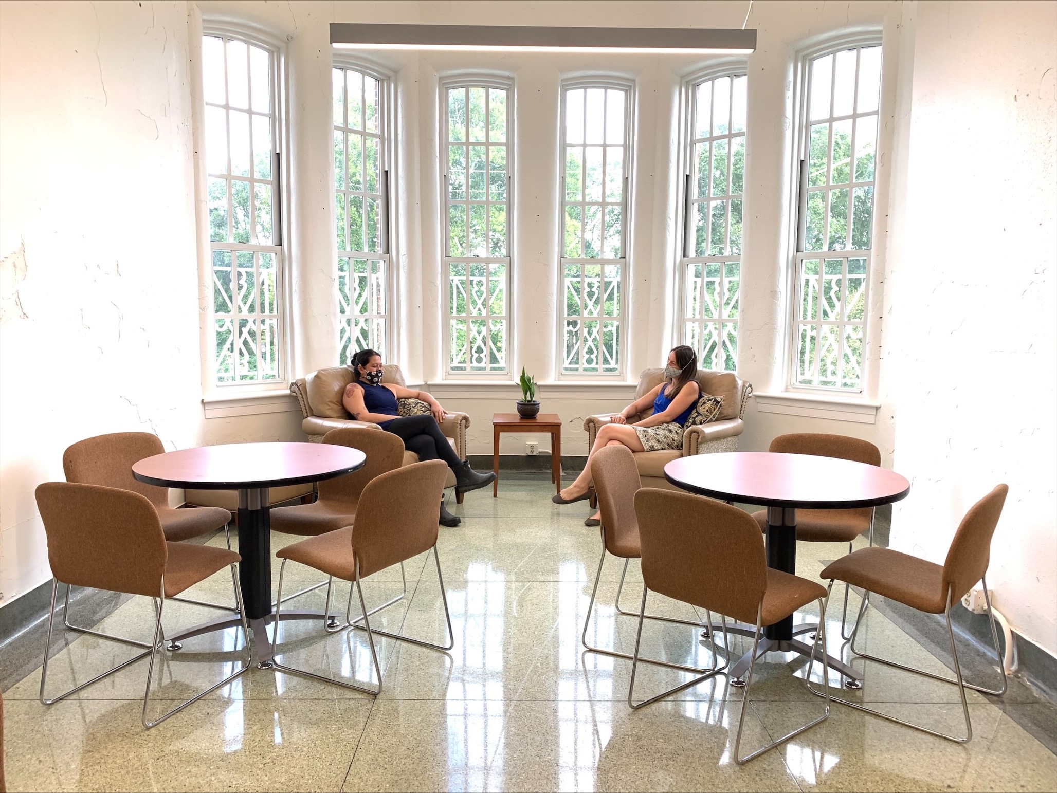 Two women sit in armchairs by windows, with two empty tables in front of them, each with four chairs