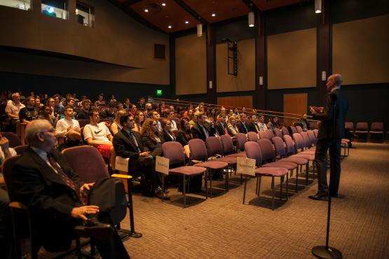 Tom De Weert speaks to students in a dimly lit Baker Center Theatre