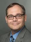 Headshot of a smiling Jesse Strycker wearing glasses and a tan suit with a blue shirt and tie.