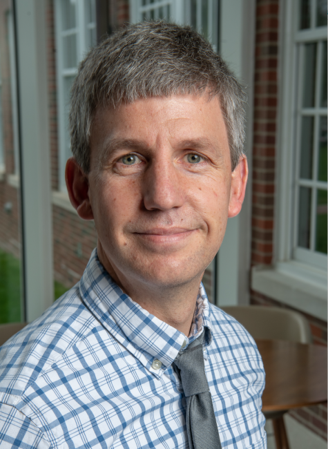 Headshot of Andy Szolosi wearing a blue plaid shirt and gray tie.