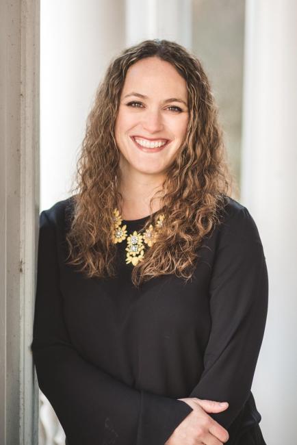 Connie Patterson leaning against a wall and smiling.  She is wearing a black top with gold accents around the neck.