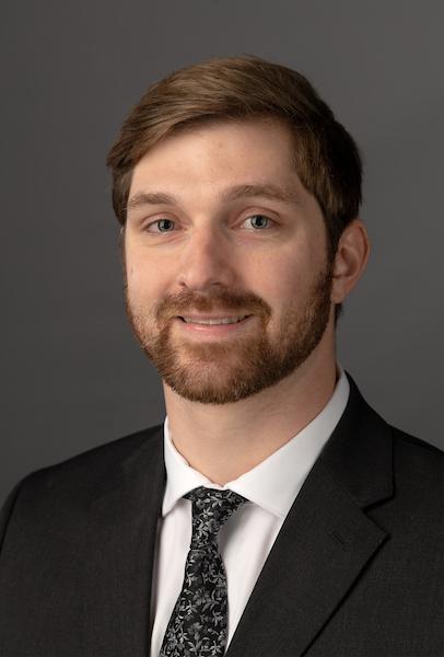 Headshot of Zac Schabel smiling wearing a black suit and tie