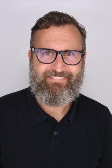 Headshot of a smiling Ryan Pigman who has a beard and moustache and wears glasses.