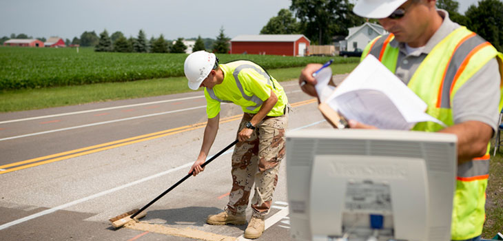 On a two-lane road next to a farm, a worker uses a push broom to spread a mud-like substance on the road surface while a second worker consults some papers