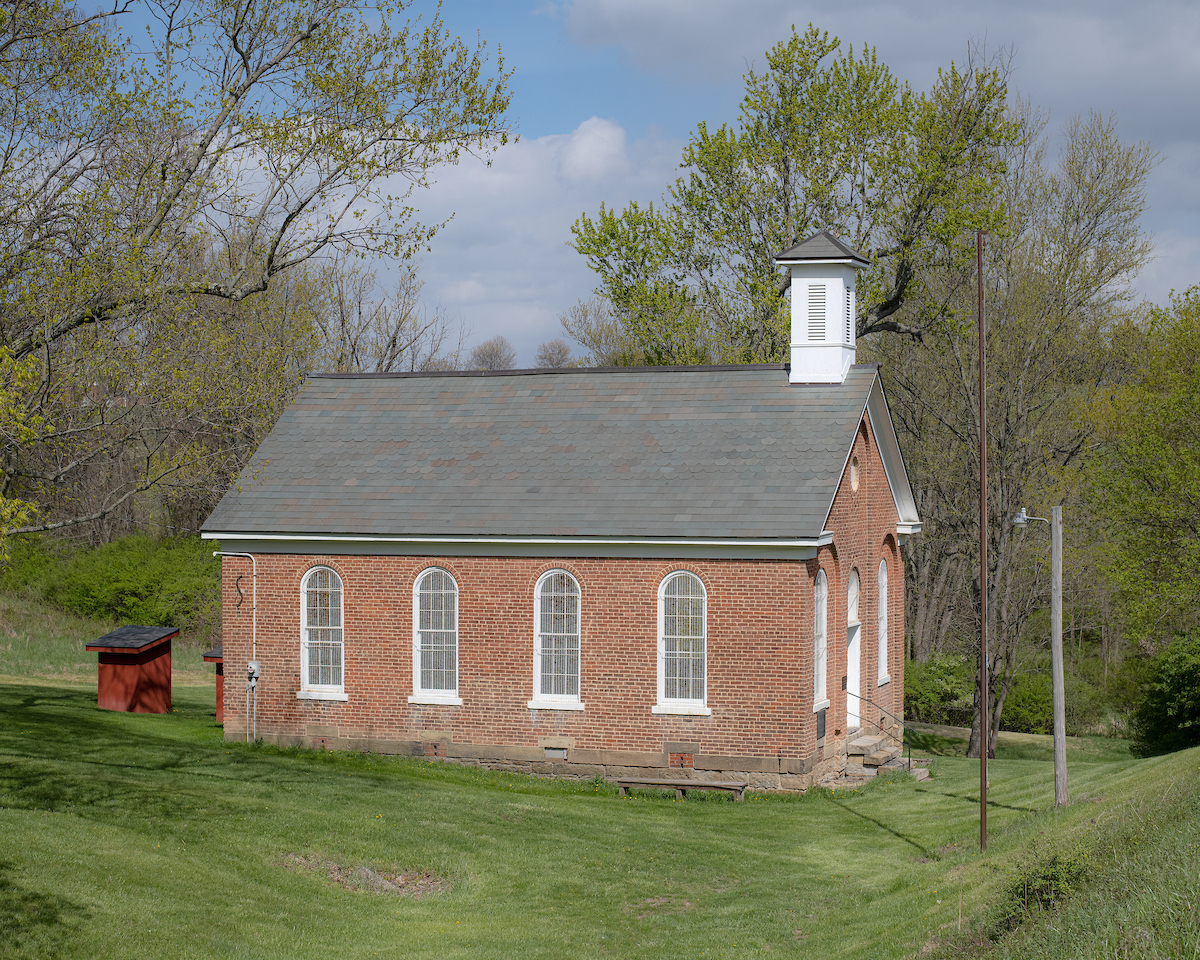 A small, historical brick schoolhouse with a few tall windows. it has a small white steeple and a grey tiled roof.
