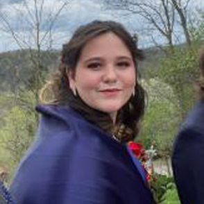 Molly Murphy looks at the camera, they are wearing a dark blue coat, and hills can be seen in the distance.