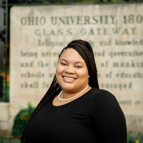 Darian Andrews smiles in front of the Ohio University Class Gateway, she wears a black shirt and a pearl necklace.