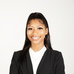 Demi Ward smiles at the camera in front of a white wall, she wears silver hoop earrings and a black blazer with a white shirt underneath.