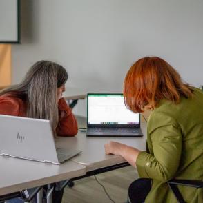 two people working at a table, with two laptops. The people are leaned over, examining one the content on one of the laptop screens.