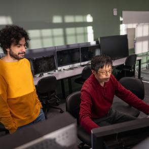 two males in front of computers, one is standing, one is sitting