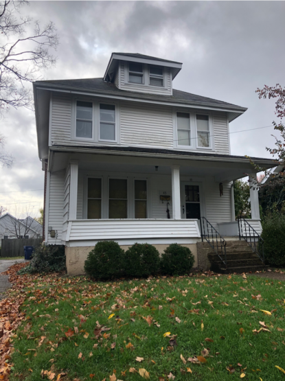 Two-story house with dormer window on attic, wood or vinyl siding, front porch and small front lawn, in a residential neighborhood