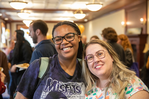 Two people stand close together side by side, smiling at the camera