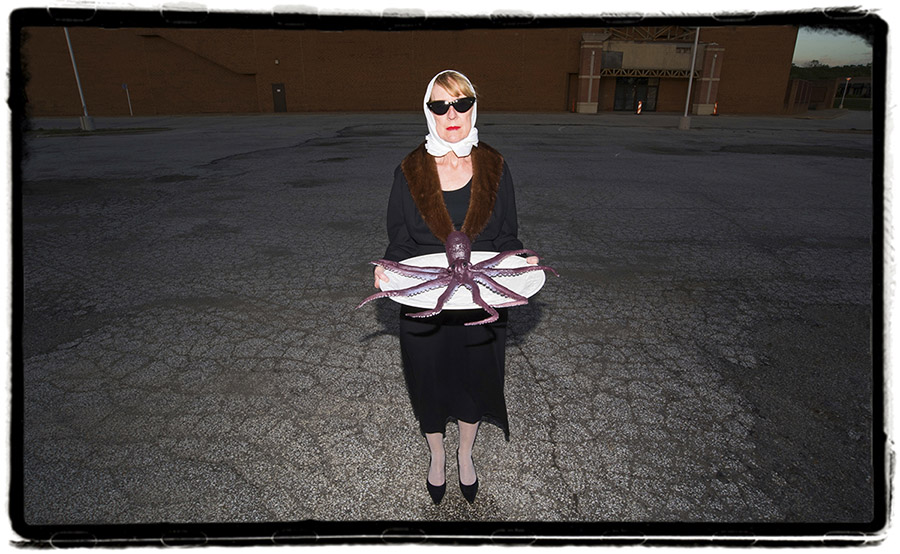 a photo of a white woman at dusk wearing sunglasses, a white kerchief covering her blonde hair, a black coat with brown fur lining and black heels; she is holding a large white plate with a purple plastic octopus on it