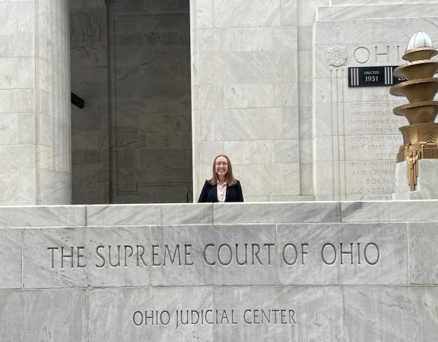 Kirsten Thomas poses behind a marble wall that reads "Supreme Court of Ohio" with below it "Ohio Judicial Center"