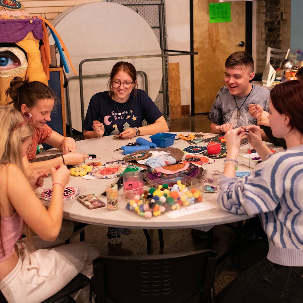 Students sit at a table making crafts together while talking and laughing.