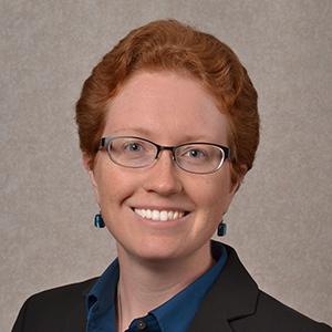 Headshot of Rachel Mauk - Smiling woman wearing glasses with short read hair and dark suit jacket