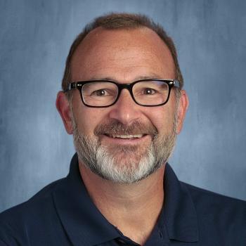 Professional Headshot of Kevin Davis, smiling man with a gray and brown beard and short hair wearing a navy blue polo shirt. He is wearing black framed glasses and the background is textured blue backdrop.