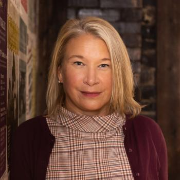 Professional Headshot of Laurie McKnight. Woman smiling at the camera with chin length blond hair, plaid blouse and cranberry sweater. The background is a postered coverd wall to her right and brick wall behind her.