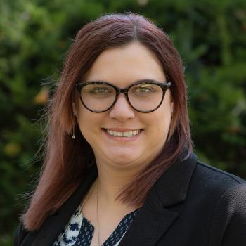 Headshot of Melanie Quokka. Woman smiles at the camera. Her hair is brunette and shoulder length and is wearing dark rimmed glasses. She is wearing a black suit jacket with a black and white patterned blouse. She is outside with a backdrop green, leafy trees. 