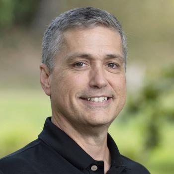 Headshot of Richard Carr. A man smiles at the camera, he has short gray hair and is wearing a black collared shirt. The background is outdoors with unfocused trees and landscaping.