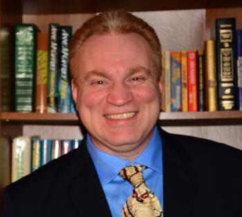 Professional Headshot of John Woodard. Smiling man with short red hair, wearing a navy suit jacket, mid blue collared dress shirt, fastened with a patterned yellow tie. He is standing in front of a wooden bookshelf with with books.