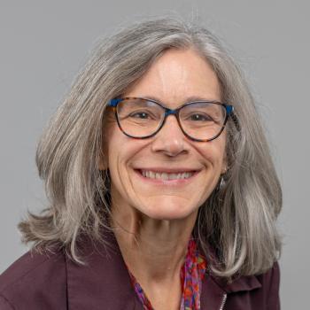 Margaret Hutzel Headshot. Woman smiles at the camera with dark rimmed glasses, shoulder length gray hair and a purple jacket and a multi colored gray backdrop. 