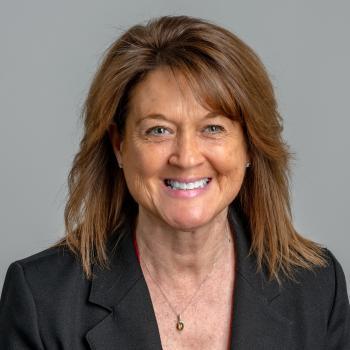 Victoria Buck Headshot. A woman smiles at the camera with shoulder length brunette hair. She is wearing a black suit jacket and the backdrop is a medium gray. 