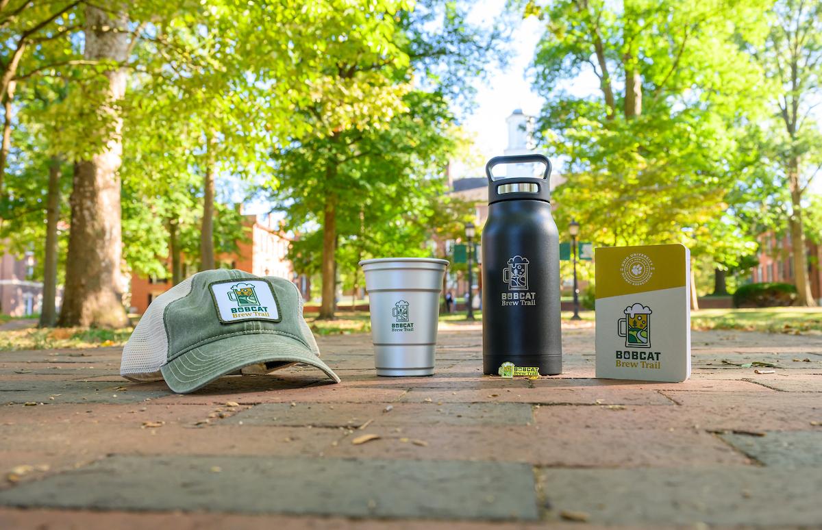 Brew Trail swag sitting on brick pathway, hat, metal cup, growler, and notebook.