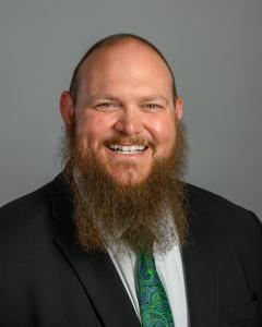 Head shot of Tyler Grubbs in front of a gray background wearing a black suit and green tie. 