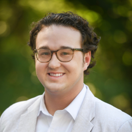 Headshot of Charlie Knox in a light suit smiling in front of greenery in the background. 