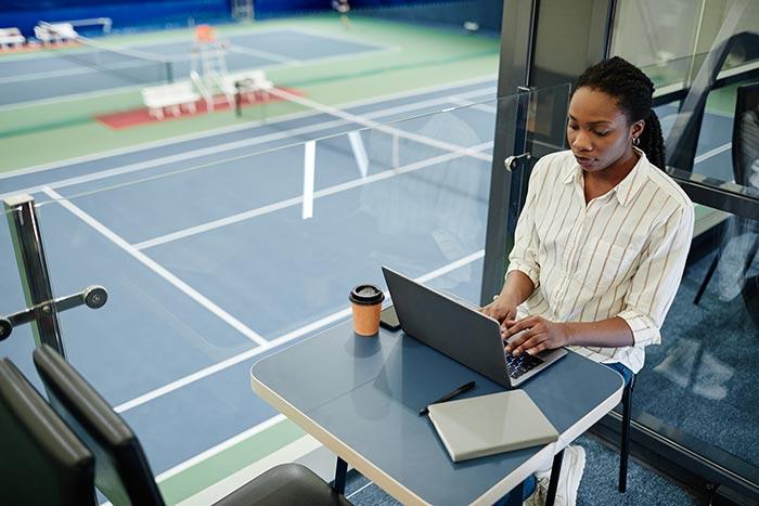 Sports manager working at her laptop on a table by window overlooking tennis courts