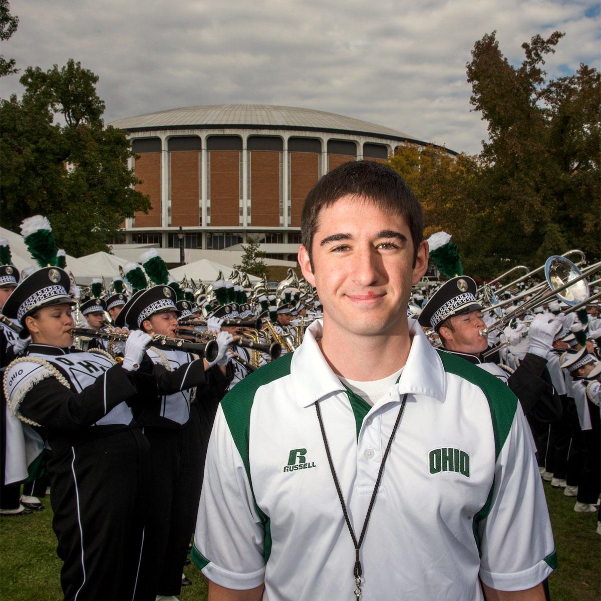 A student wearing an Ohio University photo smiles as the Marching 110 plays music behind him.