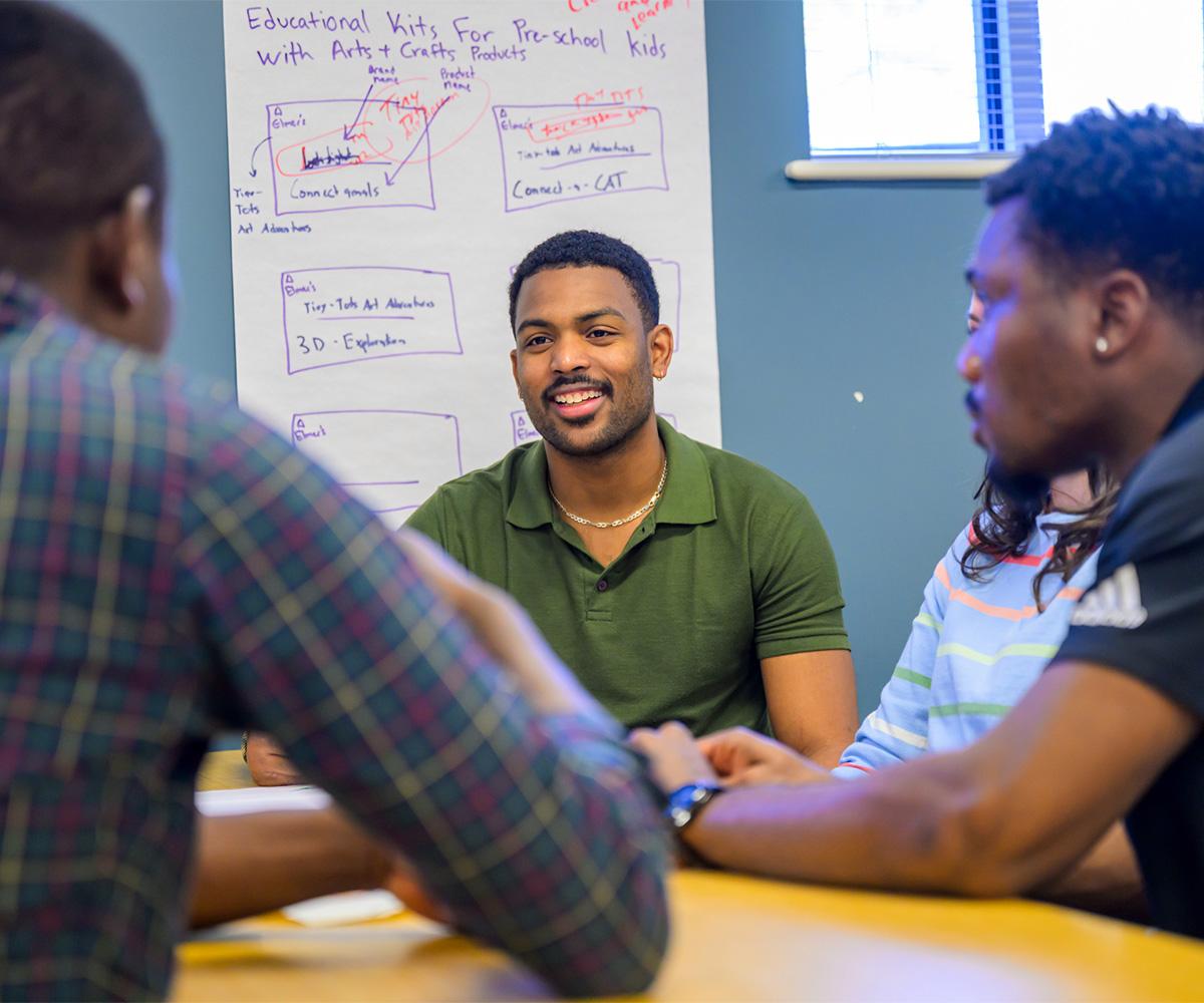 Students talk and laugh together while sitting at a table in a classroom.