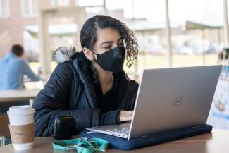Lady wearing black puffer jacket and wearing a black face mask sitting at table with open laptop