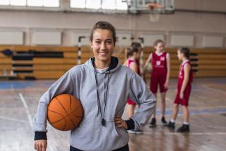 Woman smiling wearing a gray sweatshirt holding basketball in front of girls in red jerseys