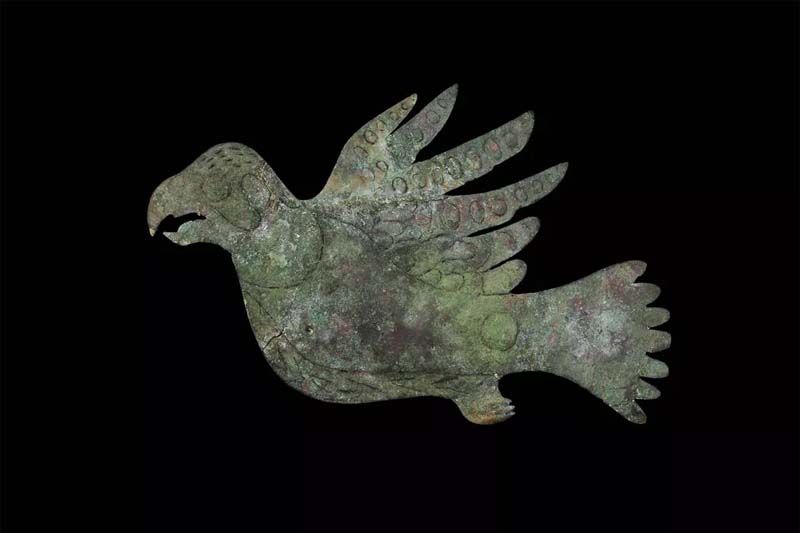 A light green-colored cutout of a bird with head to the left, wings on top, feet on bottom and tail to the right on a black background A copper effigy depicting a peregrine falcon excavated from the Mound City Group unit of Hopewell Culture National Historical Park.