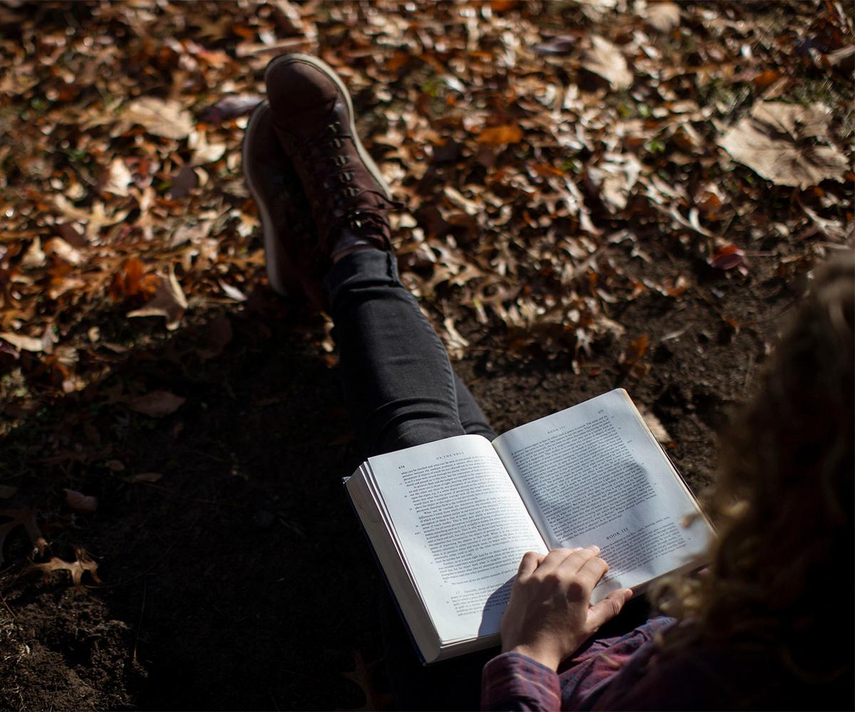 A student sits on the ground in some leaves and reads a book.