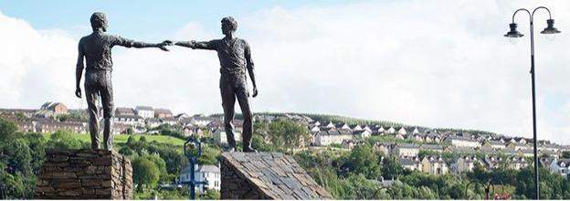 Two statues of men on brick walls reaching one arm each out towards each other