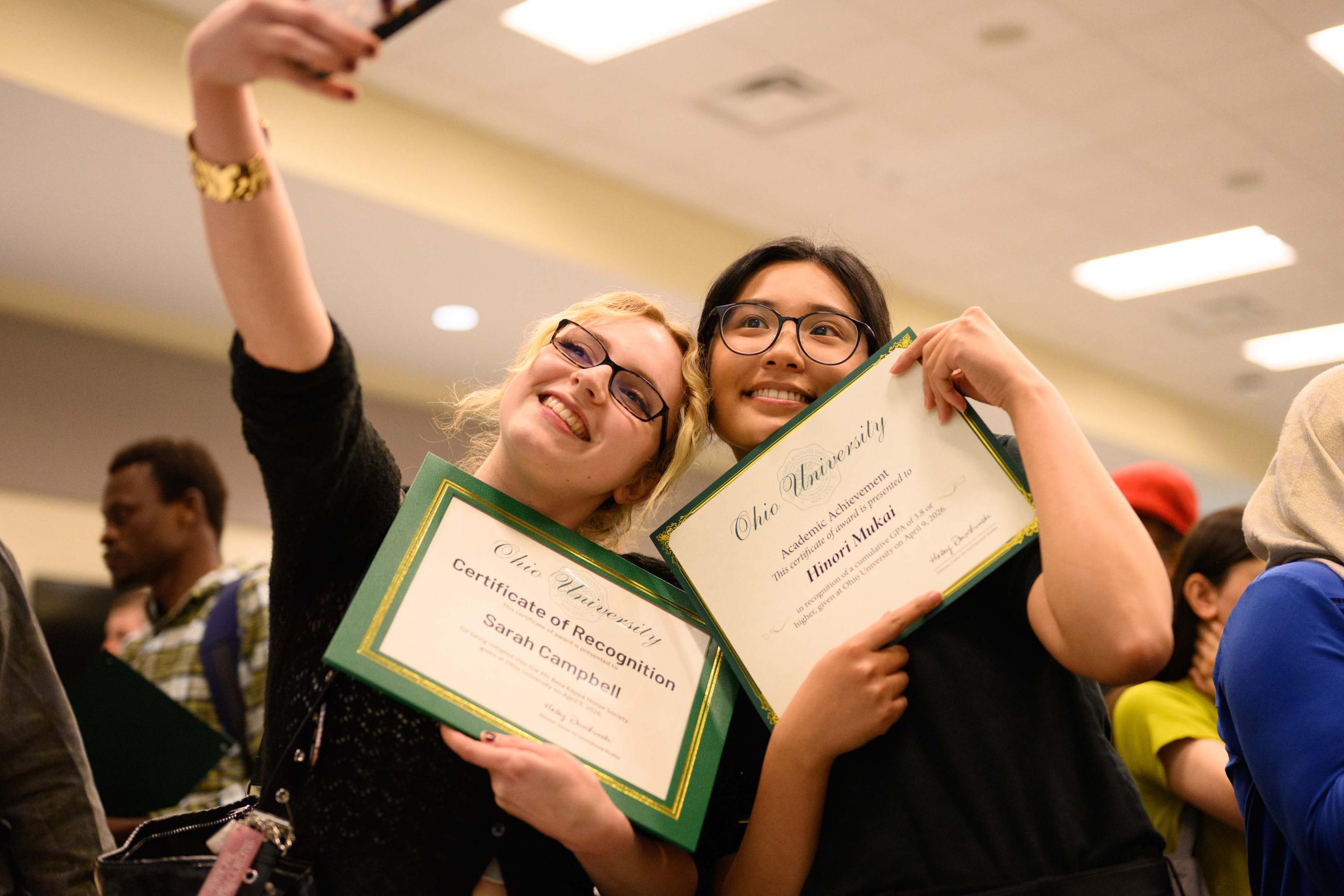 Two female students smiling for a selfie with their certificates.