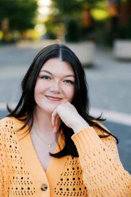 A headshot of Kaitlyn Ivey, she is smiling and resting her head on her fist, wearing a light orange cardigan