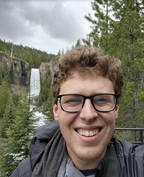 A selfie of Tyler in the forest, with a waterfall behind him.