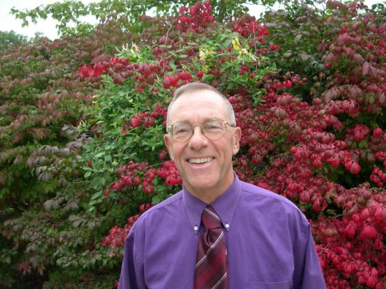 Douglas Futuyma standing in front of red and green bushes, wearing a purple dress shirt and red tie