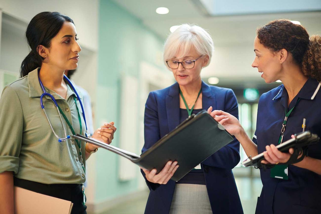 Three women talking in hallway with one holding a portfolio and one wearing a stethoscope
