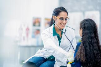 Smiling woman in white jacket and green shirt holding stethoscope up to chest of little girl