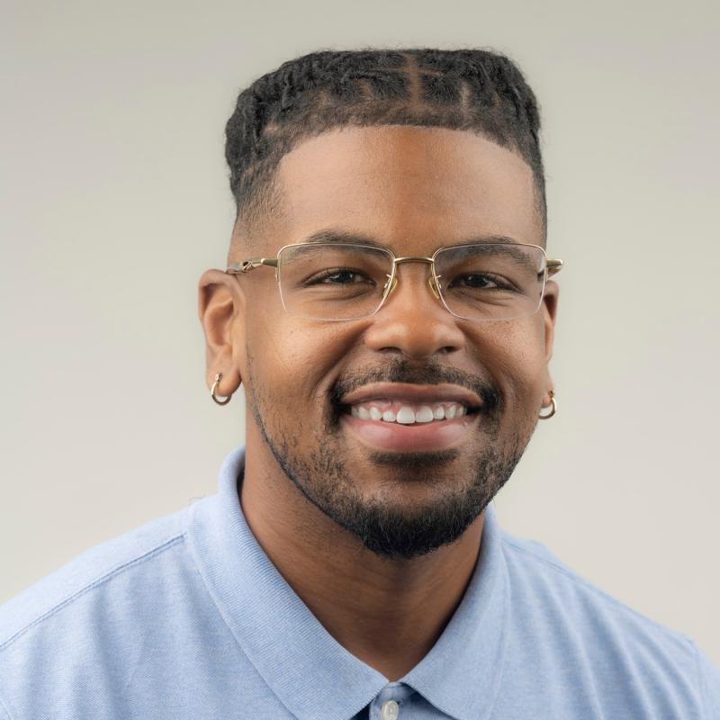 Headshot of James T. Jackson wearing a blue collared shirt and glasses without rims. Also has box braids and two small gold hoop earrings
