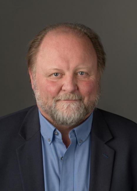 Headshot of a smiling and bearded Ken Bowald wearing a dark suit jacket and blue shirt.