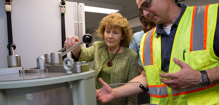A man in a reflective work vest and a woman in business attire examine a piece of equipment like a metal cabinet with protruding levers